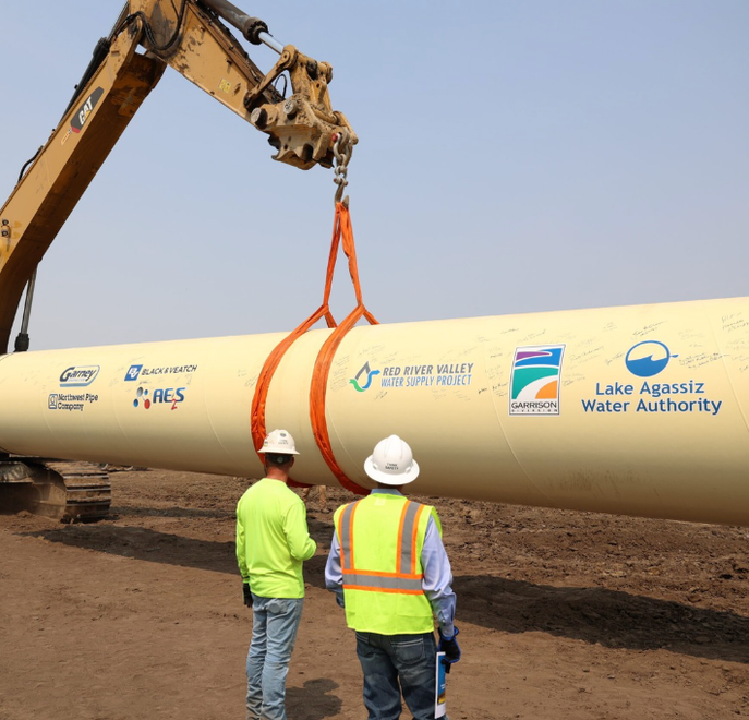 Two workers standby as a crane lifts a section of pipeline.