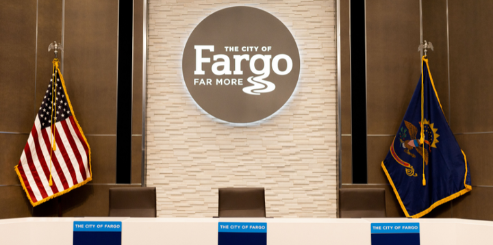 A City of Fargo emblem, the United States flag and North Dakota flag are seen behind the Fargo City Commission dais.